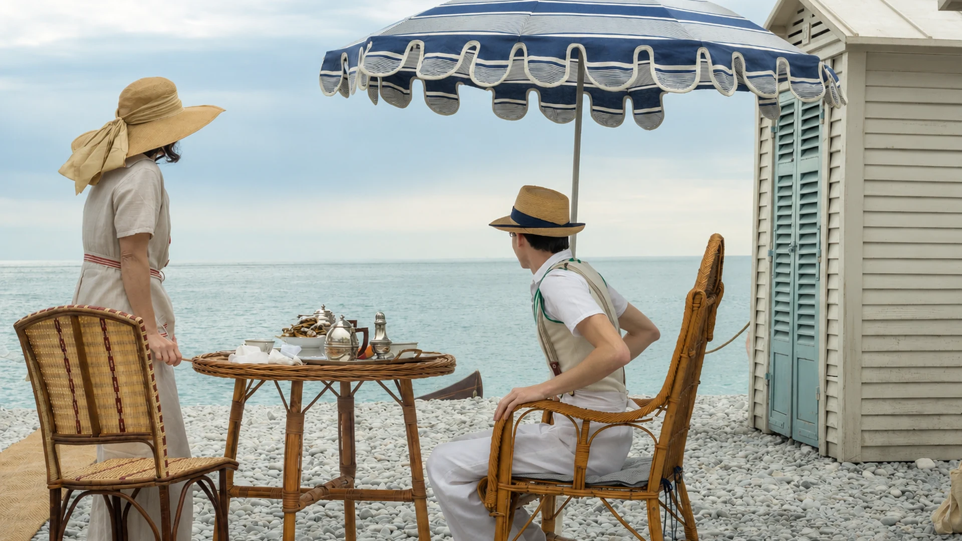 Couple bourgeois sous un parasol bleu et blanc devant une cabine de plage 1930 à Nice