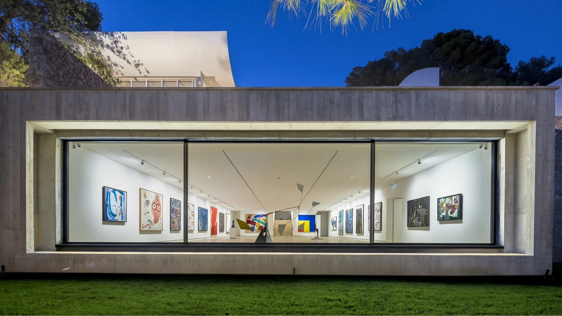 Salle d'exposition de la Fondation Maeght vue de l'extérieur en soirée : tableaux modernes éclairés derrière la baie vitrée
