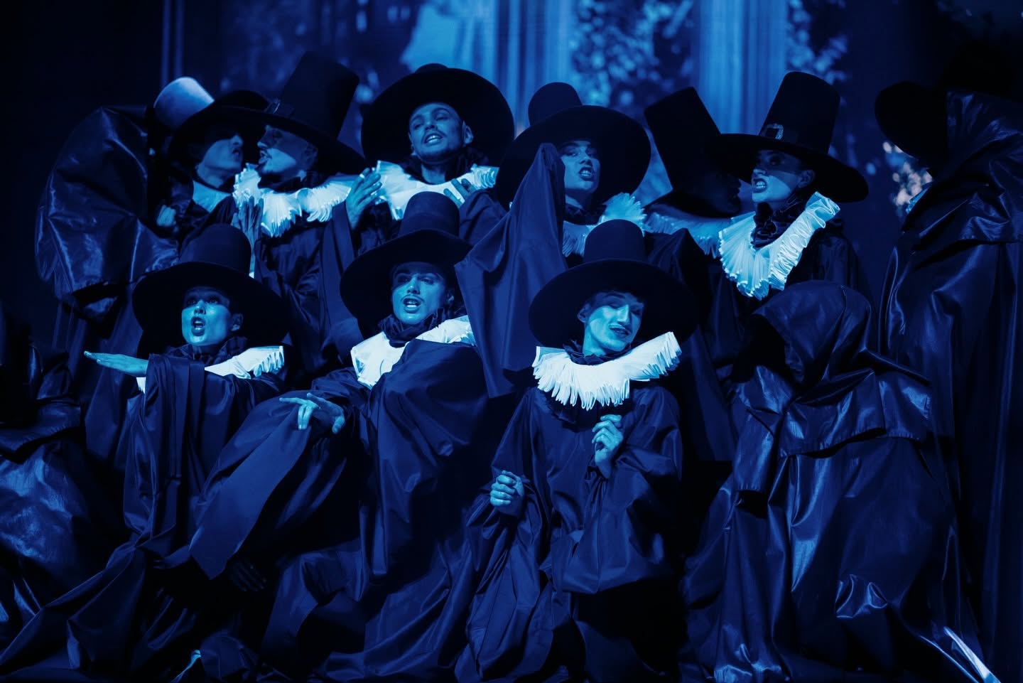 Chœur en costumes noirs et grands chapeaux baigné de bleu dans Le Roi Soleil