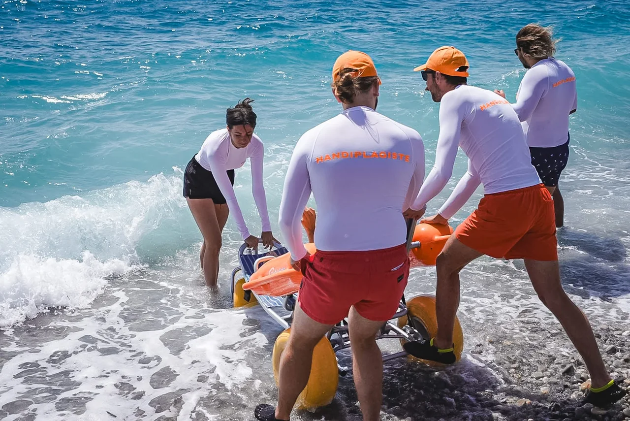 Quatre handiplagistes mettent un fauteuil tiralo à l'eau dans les vagues à Nice