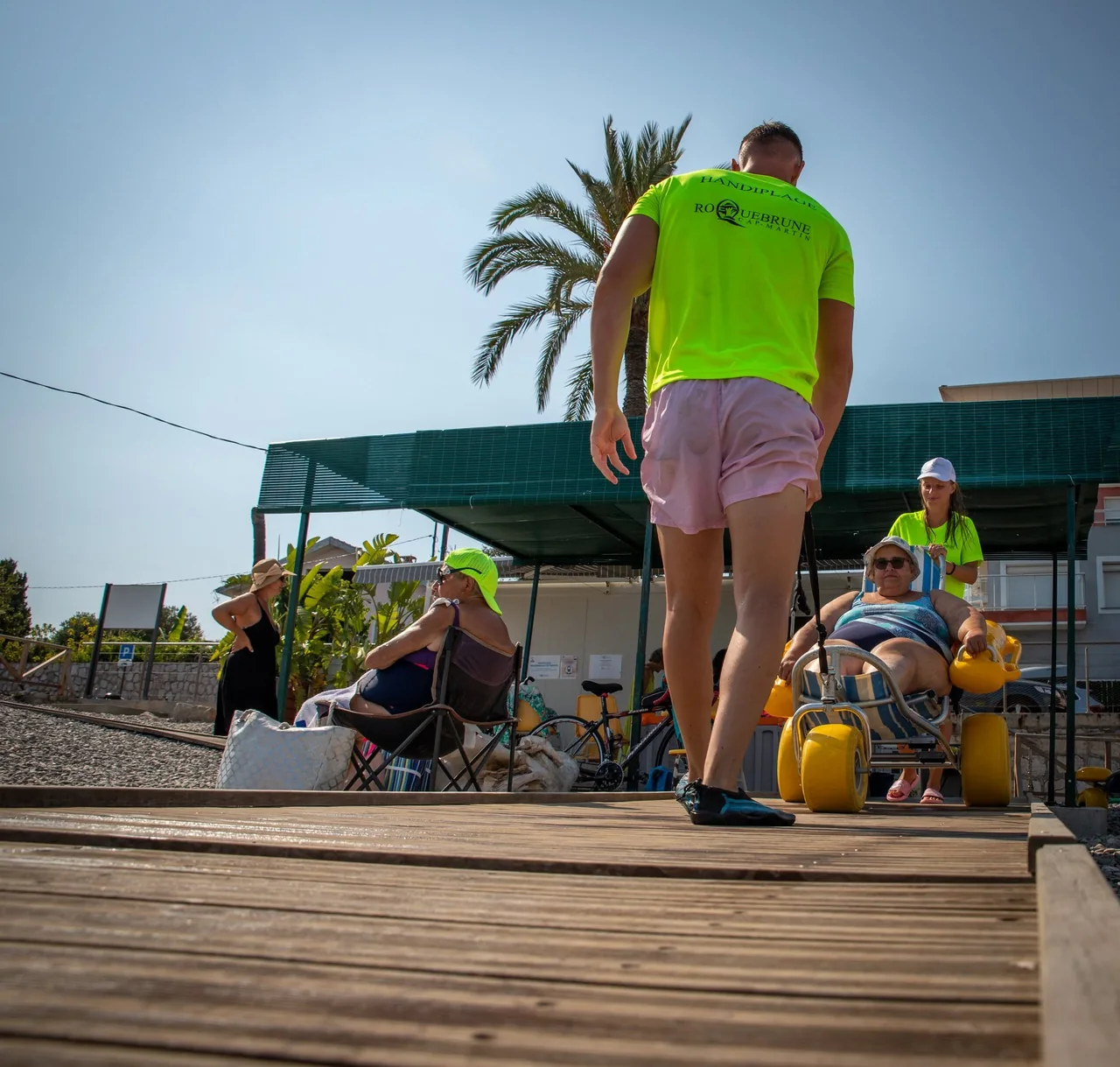 Un handiplagiste en t-shirt Roquebrune marche sur le caillebotis en bois vers la zone d'ombre de la plage Berlioz