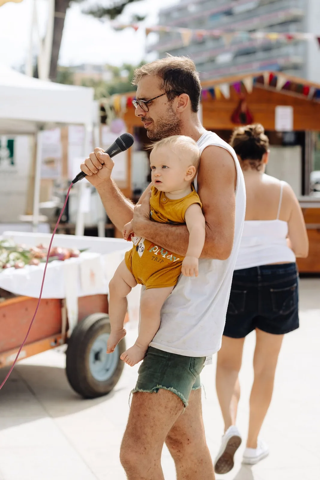 Un animateur, micro en main et bébé dans les bras, devant les stands de la fête