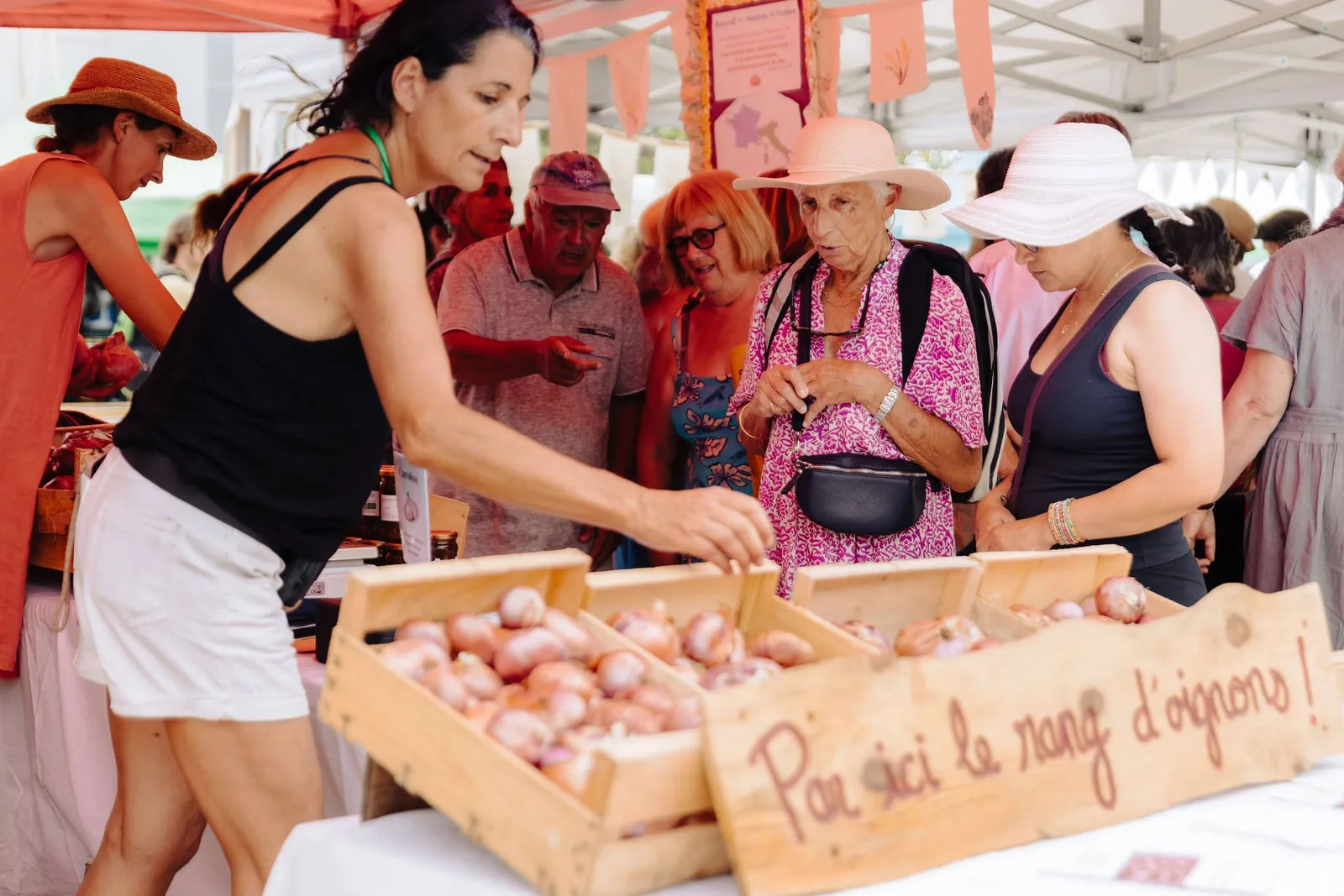 Des visiteurs se pressent autour des cagettes d’oignons roses sous un panneau « Par ici le rang d’oignons »