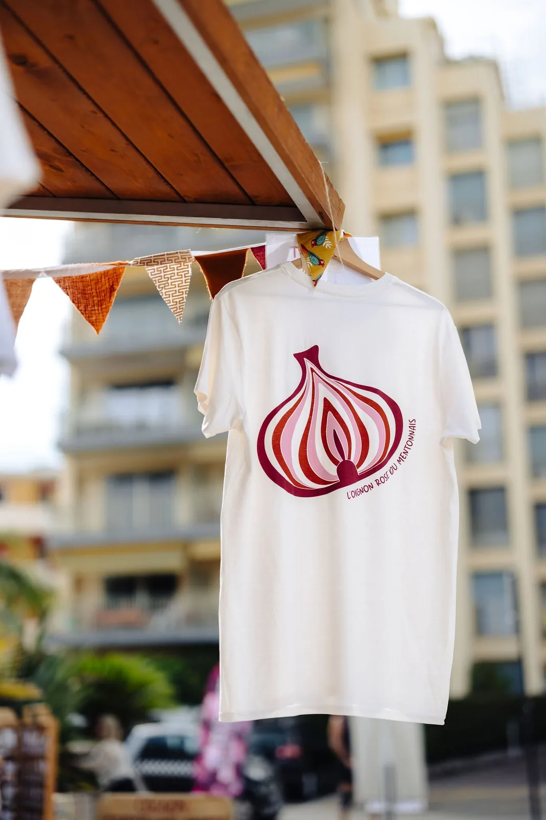 Tee-shirt blanc floqué du logo de l’oignon rose du Mentonnais, suspendu à un stand
