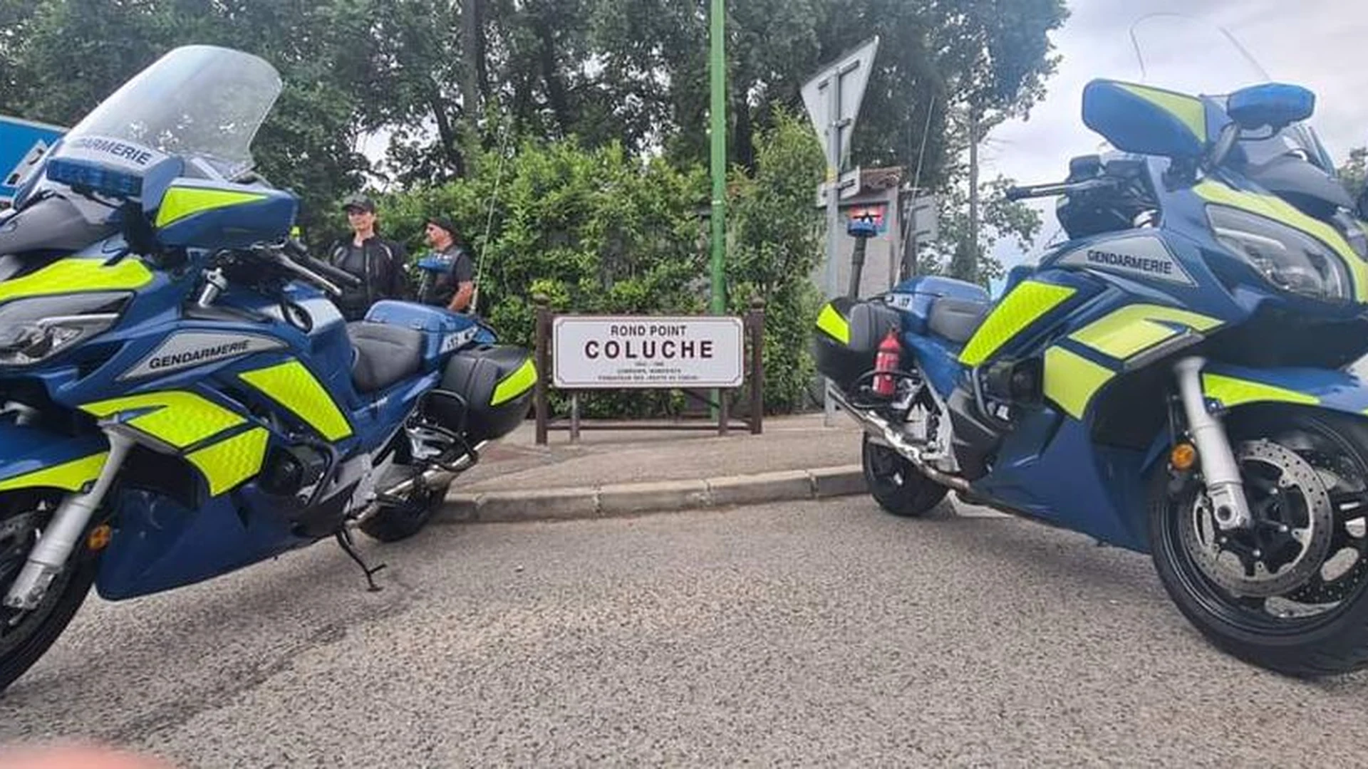Motos de gendarmerie devant le panneau Rond Point Coluche