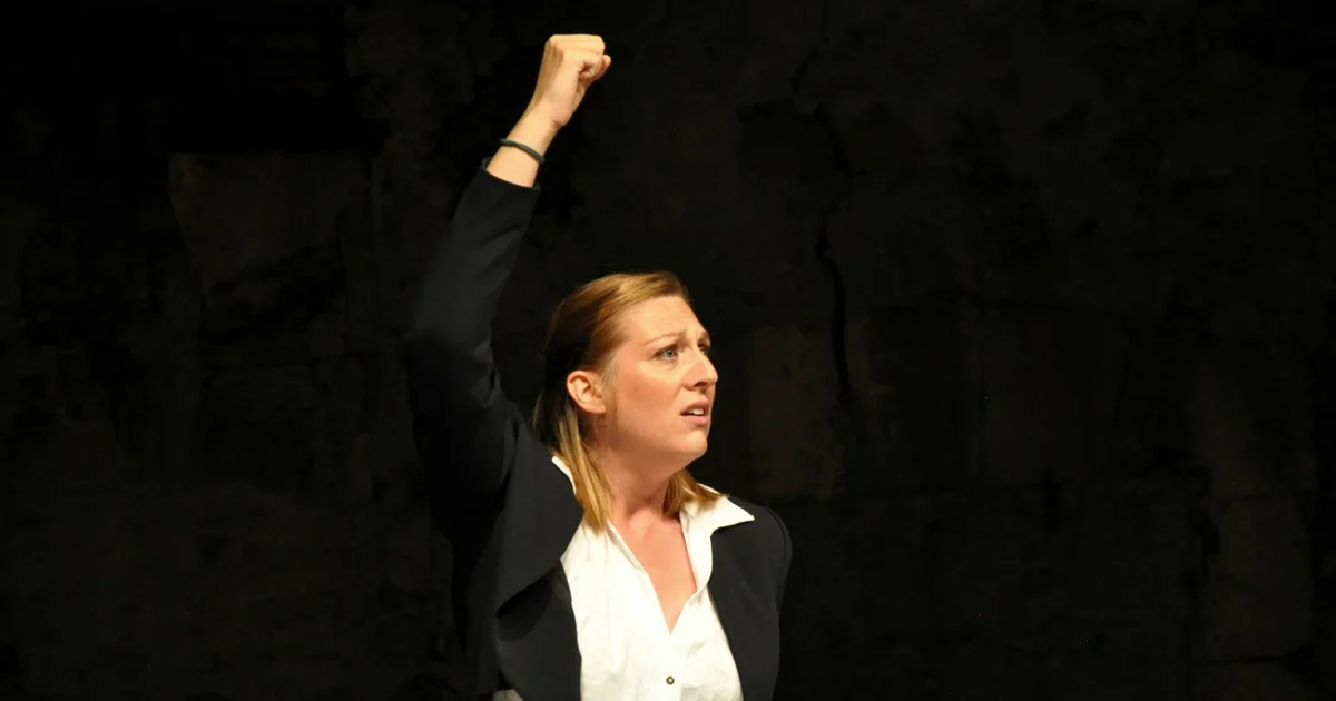Une comédienne en chemise blanche lève le poing sur fond noir