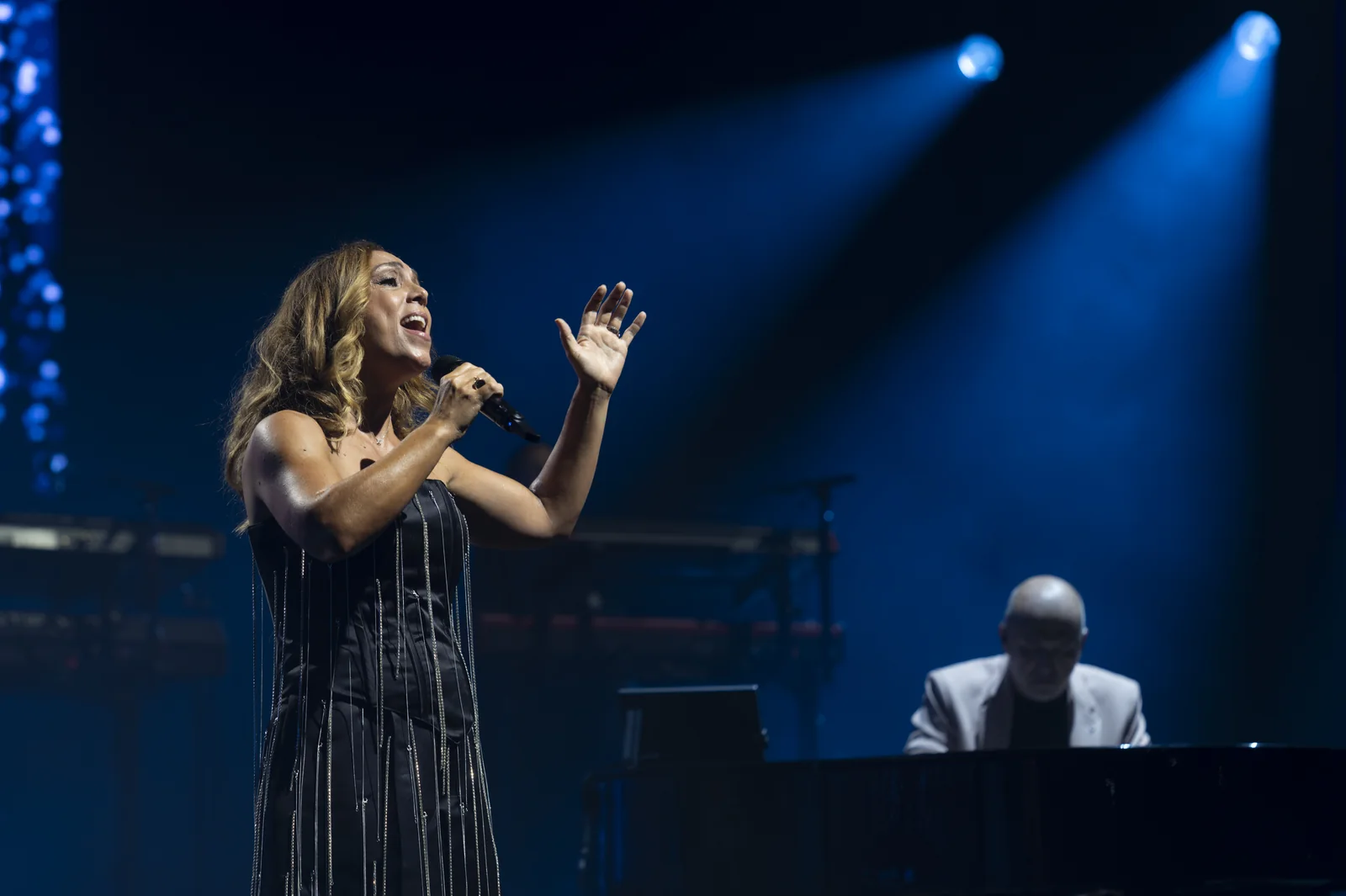 Magali Ponsada chante au micro en robe noire à franges, un pianiste dans la pénombre derrière elle