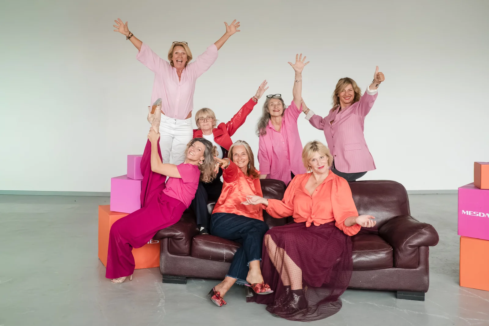 Les huit femmes de « Mesdames en vraies » autour d’un canapé de cuir, en rose, fuchsia et orange, bras levés, devant des cubes roses et orange