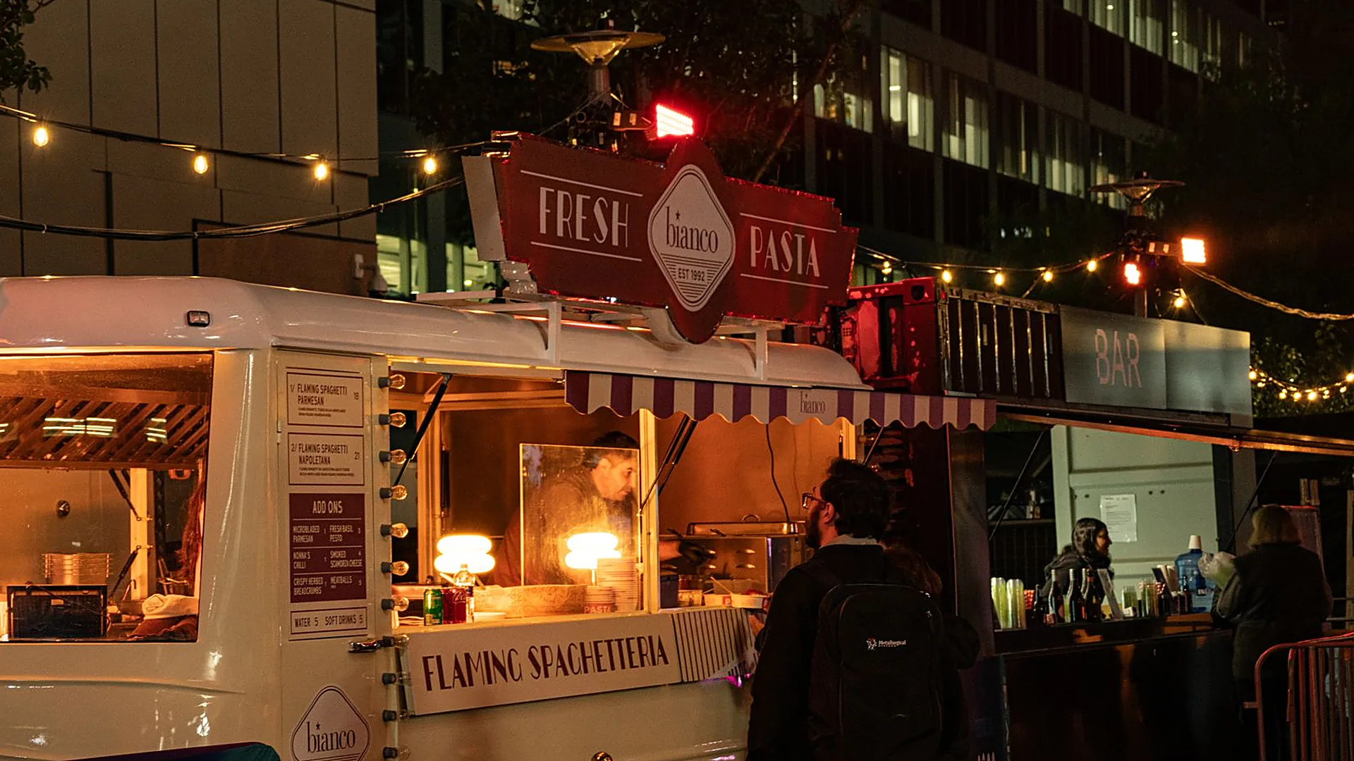 Un food truck éclairé le soir, un cuisinier au passe-plat et un client qui commande