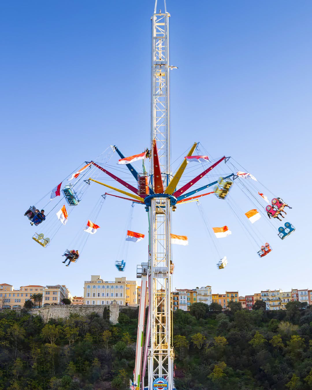 Manège de chaises volantes aux drapeaux monégasques, sous un ciel bleu, devant les façades colorées de Monaco