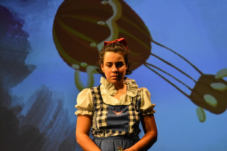 Dorothy en robe à carreaux devant un décor bleu où se dessine une montgolfière