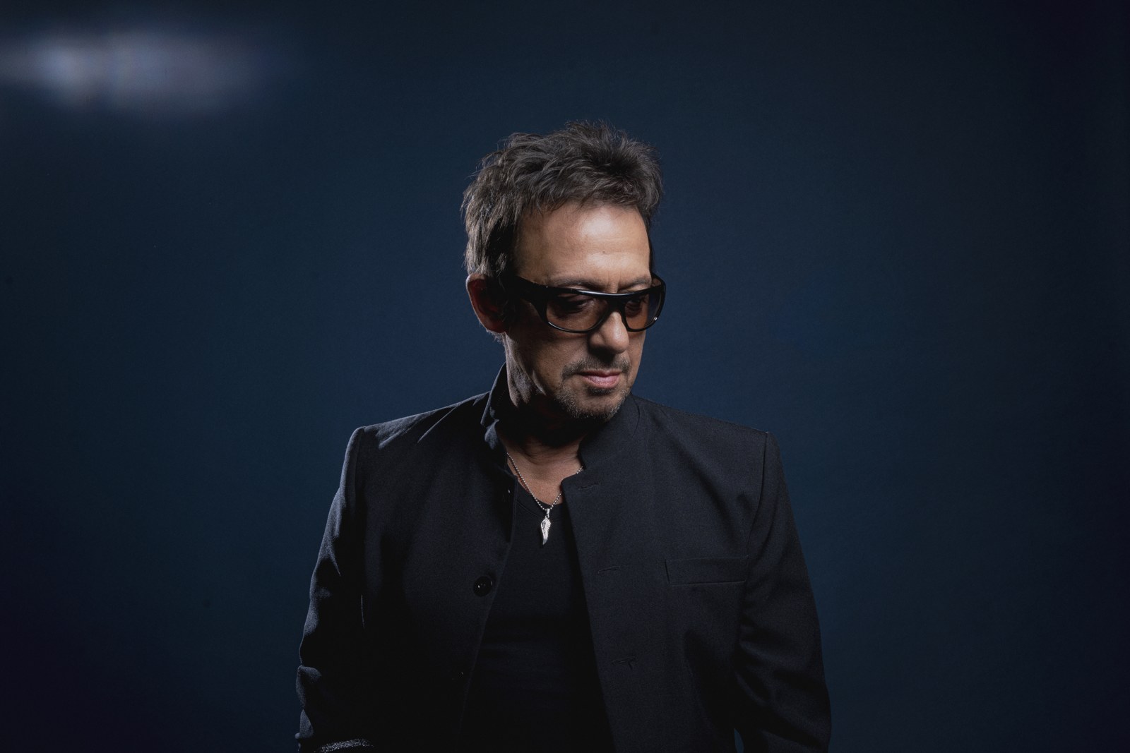 Éric Serra en veste noire et lunettes teintées, sur fond bleu nuit